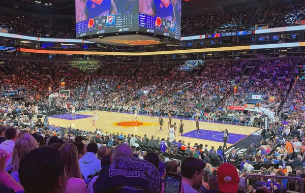Basketball game from the lower seats of a packed arena, with fans in the foreground, the court beyond and a large scoreboard overhead